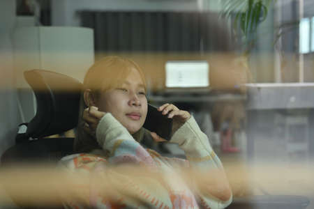 View through the window of smiling woman sitting in living room and talking on mobile phone.の写真素材