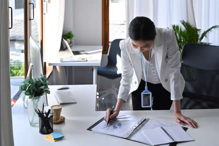Business woman standing at her offie desk and checking financial reports.
