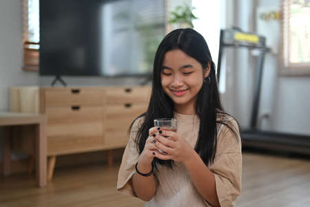 Young Asian woman holding glass of water and smiling to camera while standing in the kitchen.の写真素材