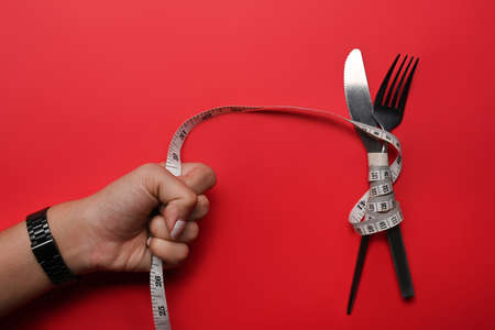 Woman hand holding measuring tape with knife and fork on red background. Loss weight and healthy diet concept.の写真素材