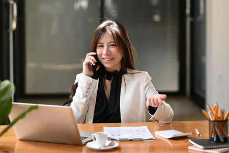 Smiling businesswoman sitting in front of laptop computer at office desk and talking on mobile...