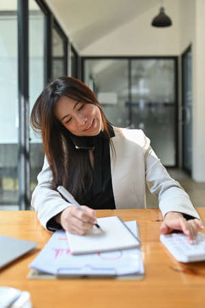 Asian businesswoman talking on mobile phone and checking reports at office desk.の写真素材