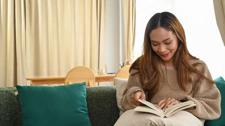 Smiling asian woman sitting on comfortable couch and reading book.