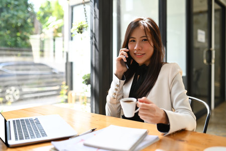 Charming businesswoman drinking coffee and talking on mobile phone.の写真素材