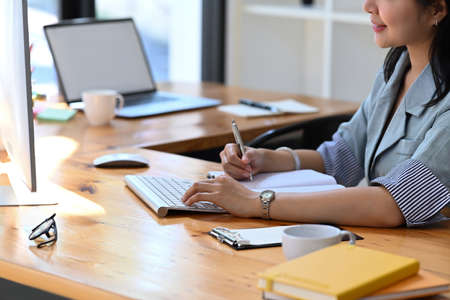 Cropped shot young female office worker working with computer and making notes on notebook.