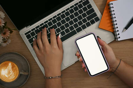 Overhead view woman holding mobile phone and using laptop computer on wooden desk.