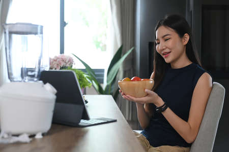 Happy woman eating healthy salad while having video call on computer tablet in the kitchen.の写真素材