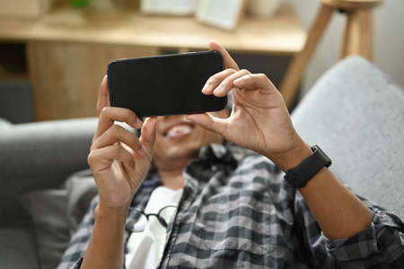 Young man lying on comfortable sofa at home and watching movie on smart phone.