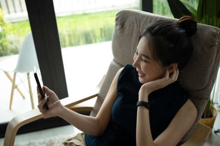 Cheerful young woman relaxing on comfortable armchair and making video call on smart phone.