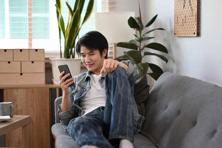 Happy young asian man lying on couch at home and using smart phone.の写真素材