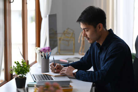 Concentrated asian man working with laptop computer and making notes on notebook.の写真素材