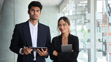 Two businesspeople in formal wear standing in modern office and looking confidently at camera.