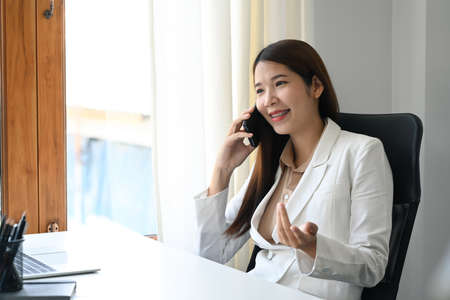 Millennial asian business woman sitting in bright office and talking on mobile phone.