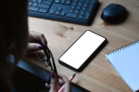 Female office worker holding eyeglasses and using smart phone on wooden desk.