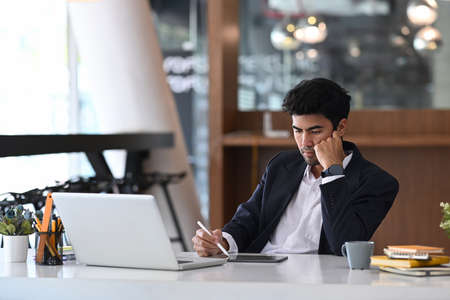 Handsome young businessman sitting at office desk behind laptop computer and searching information on digital tablet.の写真素材