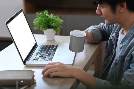 Man freelancer in casual wears drinking coffee and browsing internet with laptop at home.