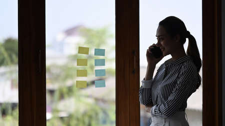 Experienced standing in a modern office interior near a large window and talking on mobile phone.
