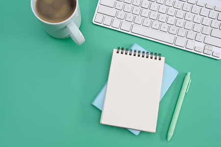 Empty notepad, coffee cup and wireless keyboard on green background. Flat lay.