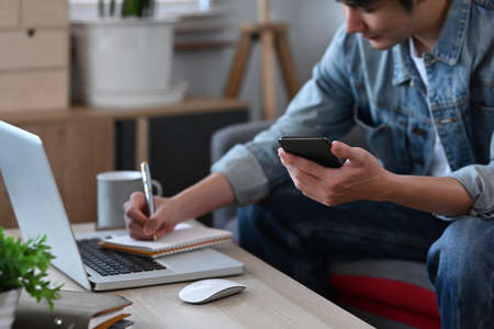 Smiling asian man holding mobile phone and making notes on notebook while working from home.