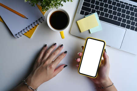 Above view woman hand holding mobile phone with blank screen on white office desk.の写真素材
