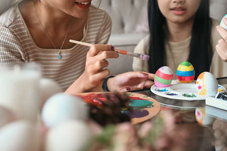 Happy young mother hands holding paintbrush helping her daughter painting Easter eggs. Easter,...
