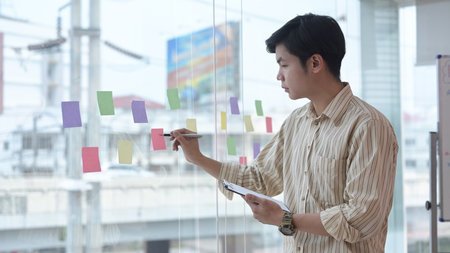 Thoughtful male employee writing information on colorful sticky notes develop business project in office.の写真素材