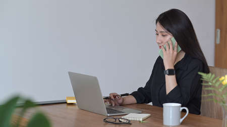 Busy female entrepreneur using laptop computer and talking on mobile phone with her business...