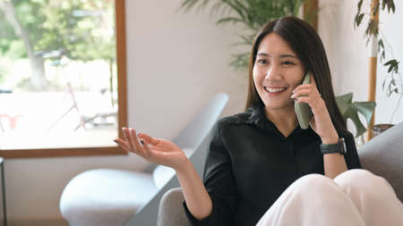 Cheerful young woman resting on couch at home and talking on cellphone.の写真素材