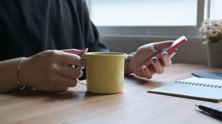 Stylish man hand holding coffee cup and using mobile phone on wooden table.の写真素材