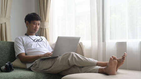 Happy asian man using laptop computer while relaxing on a comfortable couch at home.