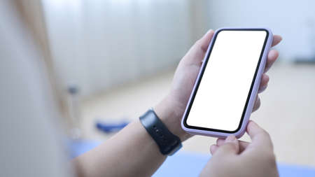 Close up view young woman sitting on yoga mat in living room and using smart phone.