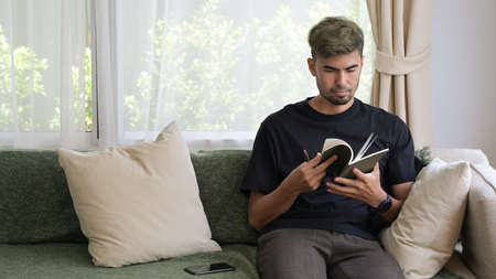 Handsome young man in casual clothes reading a book while sitting on couch at home.の写真素材