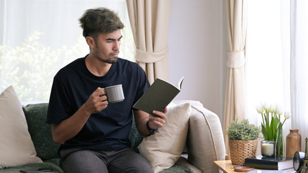 Handsome young man in casual clothes reading a book while sitting on couch at home.
