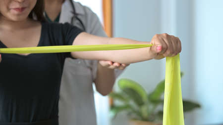 Young physiotherapist helping patient exercising arms with yellow elastic rubber at rehabilitation...