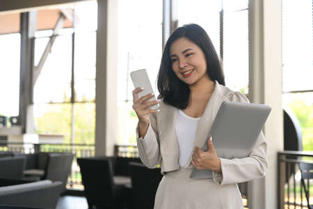 Gorgeous young businesswoman using mobile phone while standing in modern office.