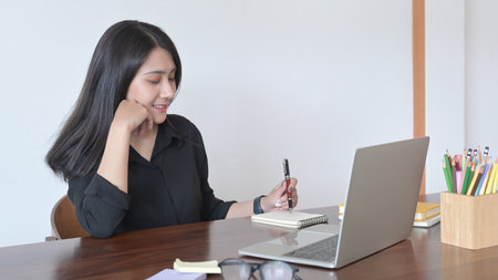 Charming  asian woman sitting in modern home office and working online on laptop.