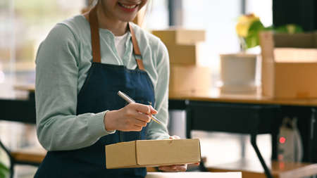 Pretty young woman writing address on cardboard box while preparing parcel boxes of product for...
