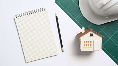 Above view blank notebook, house model and safety helmet on white table.