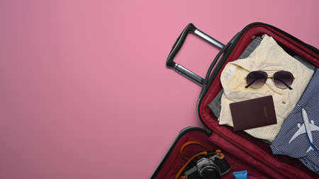 Open suitcase with sunglasses, camera, passport and clothes on pink background. Preparing for the...