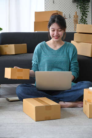 Smiling young man checking product purchase order on laptop computer and preparing parcel for delivery.の写真素材