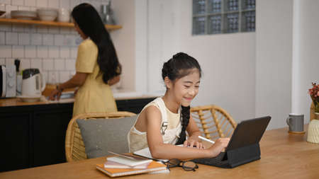 Asian girl using laptop during online class with tutor or teacher while sitting in home kitchen and...
