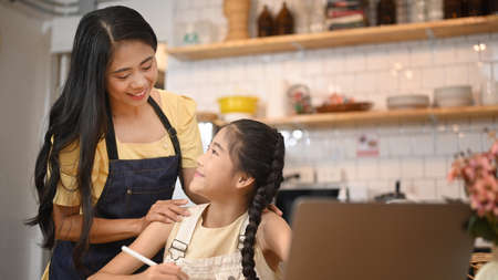 Smiling asian mother helping her little daughter with distance learning virtual distance online class on laptop computer.の写真素材