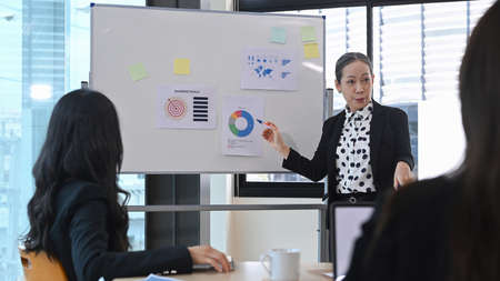 Focused senior female team leader presenting project research results to interns colleagues,...