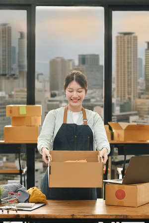 Smiling startup business owner holding cardboard and smiling to camera standing near large window at...