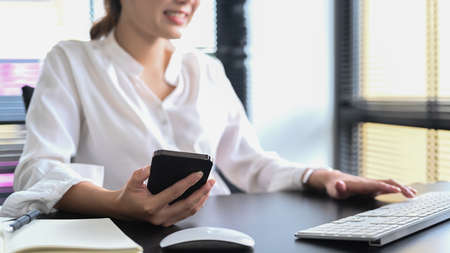 Smiling female office worker holding smart phone and typing on wireless keyboard. Cropped shot.