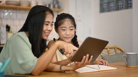 Smiling Asian mother helping her daughter doing homework, surfing internet on digital tablet at...