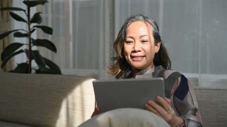 Smiling middle aged woman relaxing on comfortable sofa and browsing wireless Internet on tablet....
