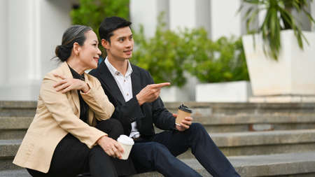 Mature businesswoman and businessman sitting on stairs of an office building and talking to each other.の写真素材
