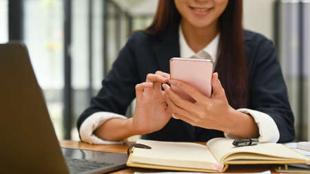 Smiling young woman sitting front of laptop in office and using smart phone.の写真素材