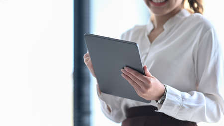 Smiling young female employee standing near window in modern office and using digital tablet.の写真素材